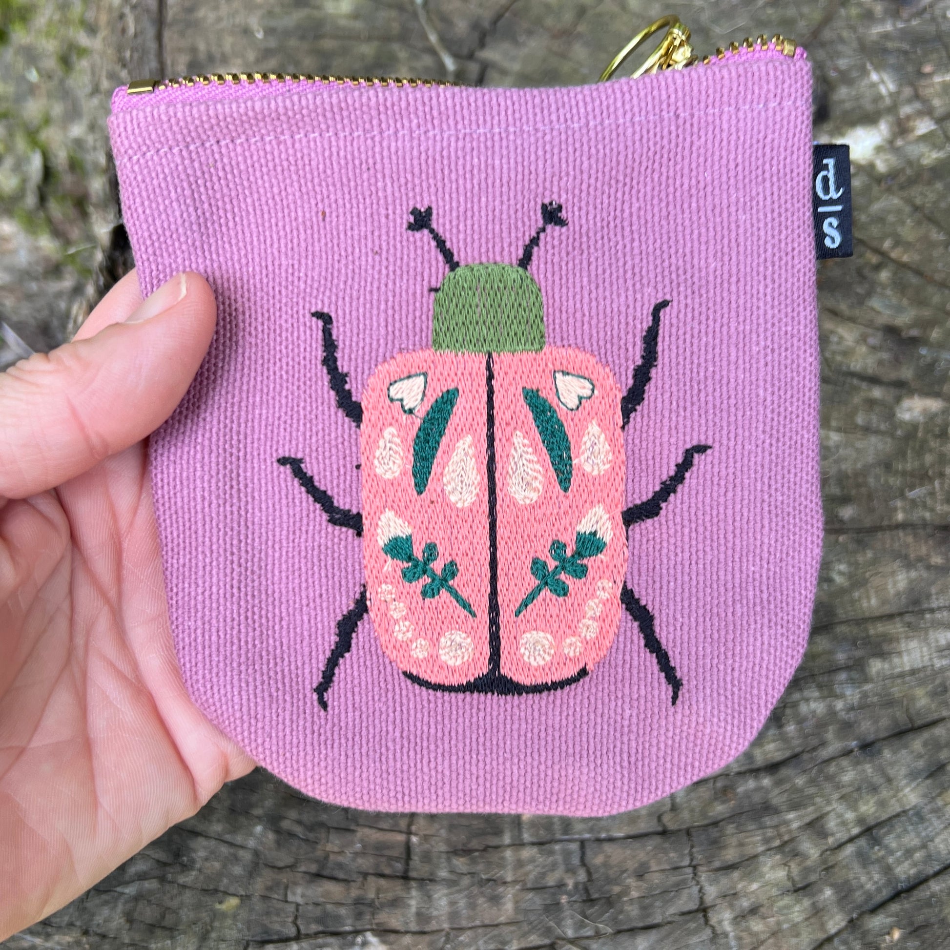 Purple pouch with a pink beetle design held by a hand on a wooden surface