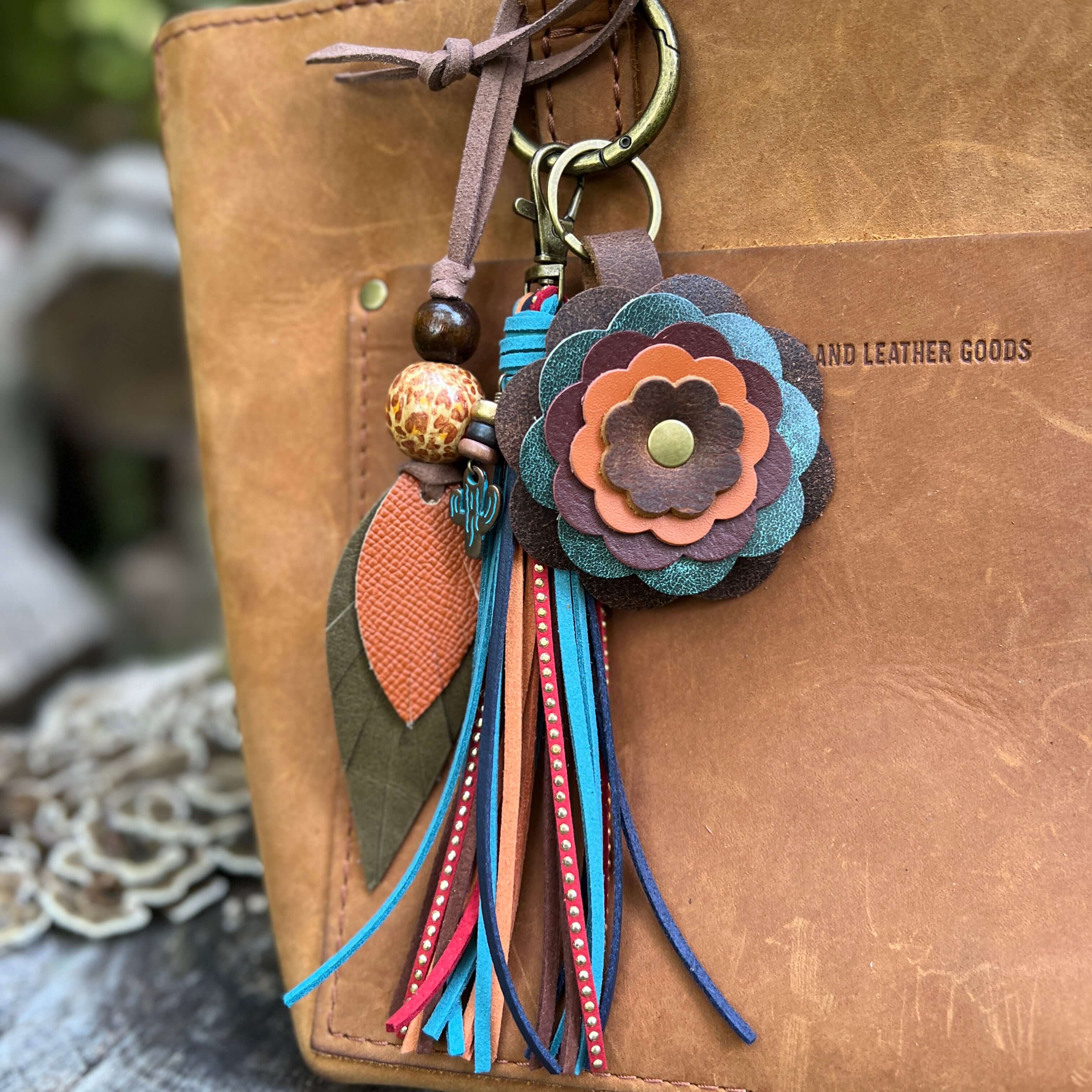 A bohemian-style leather keychain charm with layered flower petals, tassels, and decorative beads, attached to a brown leather bag. The bag has 'PORTLAND LEATHER GOODS' embossed on the side.