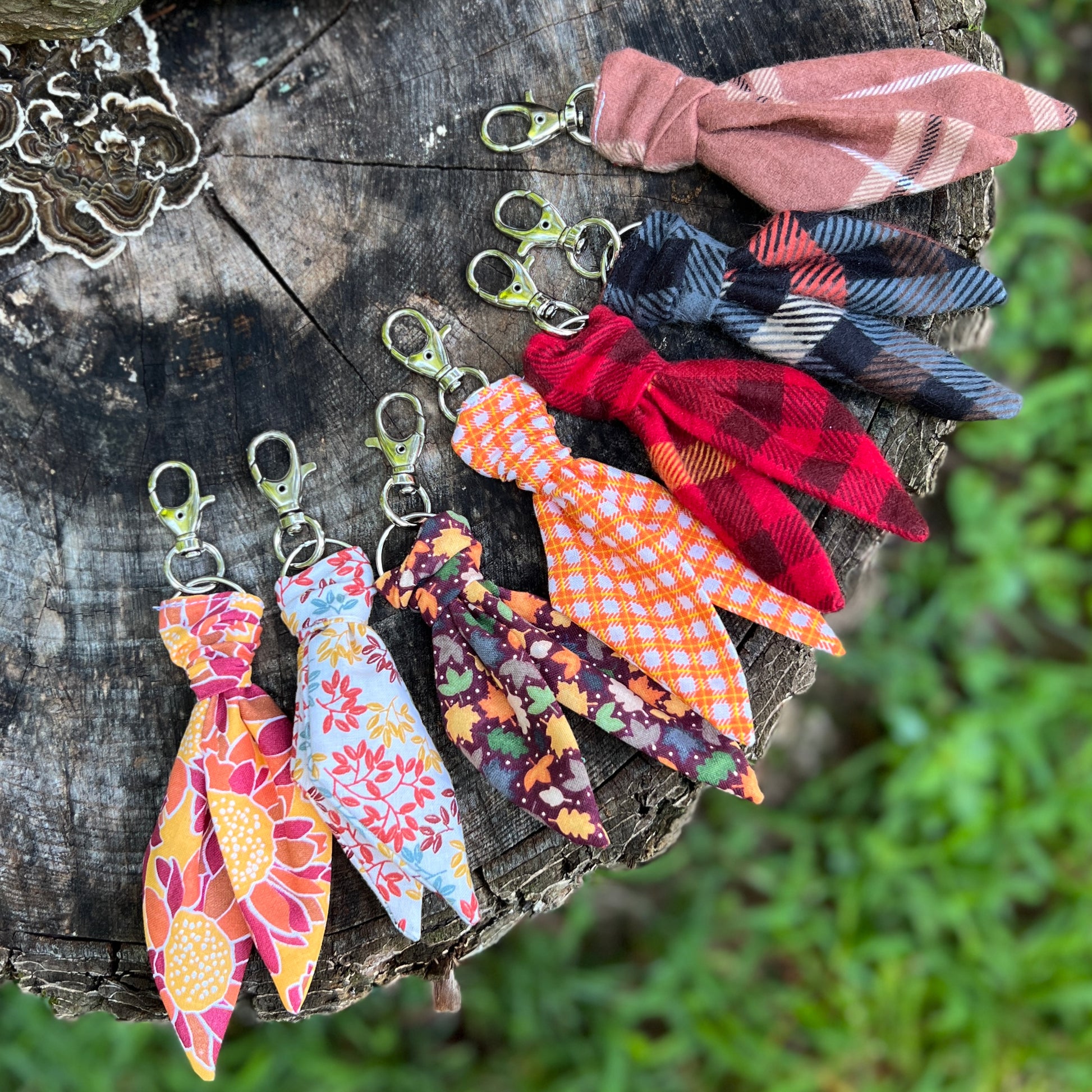 Six fabric bow keychains with different colorful patterns and prints, attached to silver metal lobster clasps, arranged on a rustic wooden log with a blurred green outdoor background.