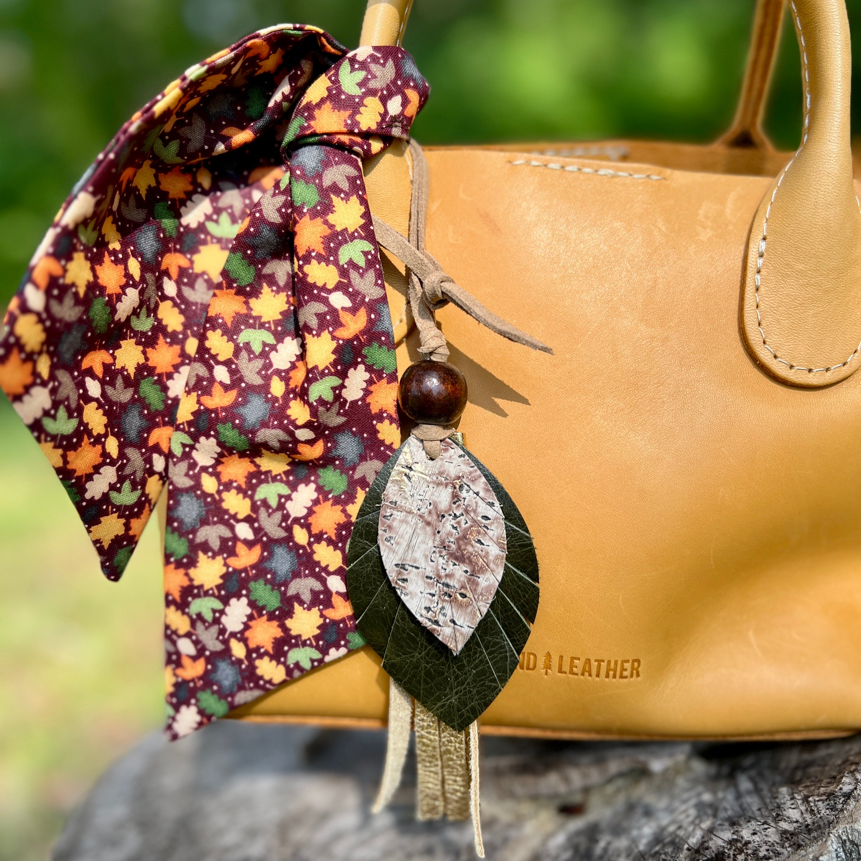 Tan leather handbag with a decorative leaf charm and colorful fabric on a natural background