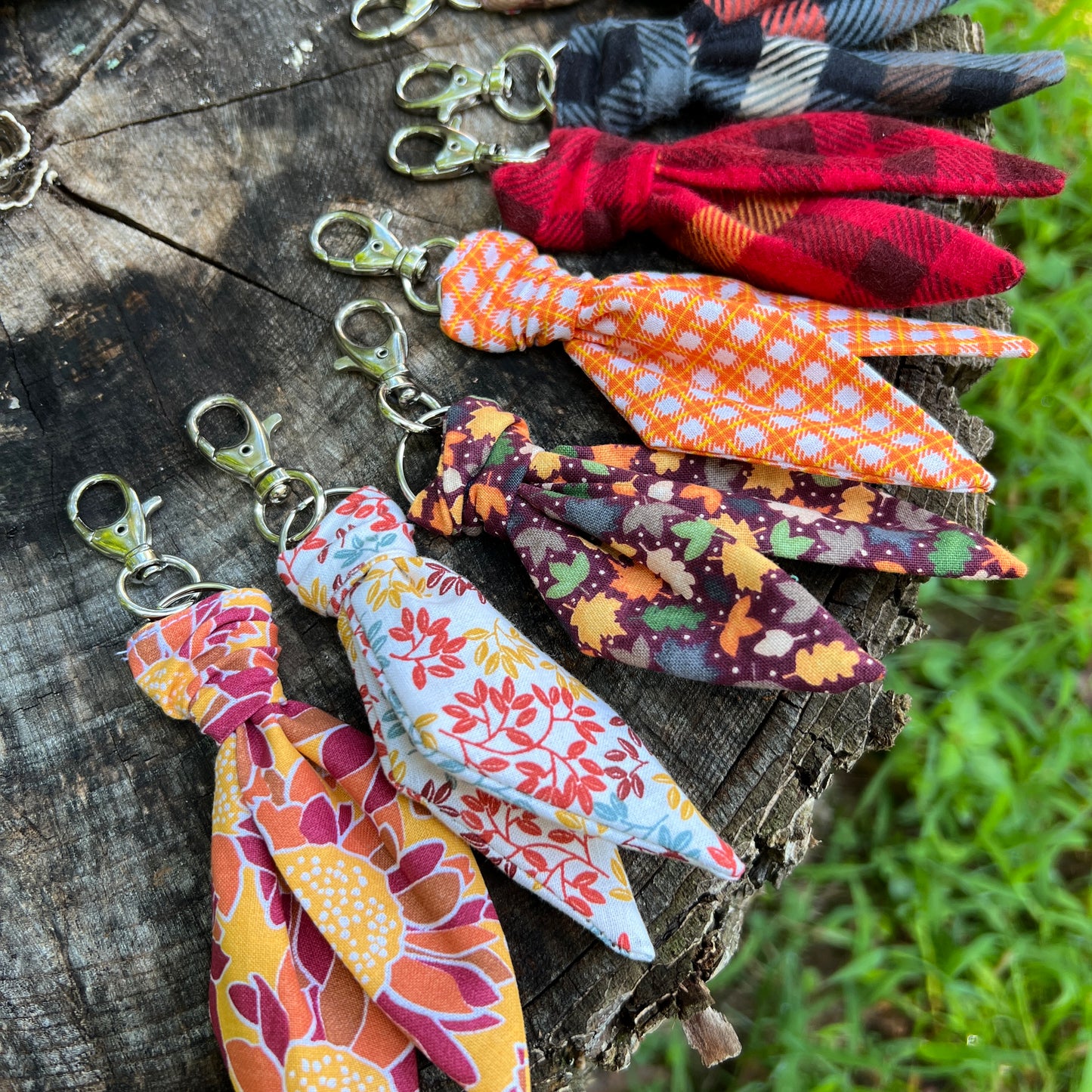 Six fabric keychains with floral and plaid patterns, attached to silver lobster clasp keys, arranged on a rustic weathered wood log with green grass in the background.
