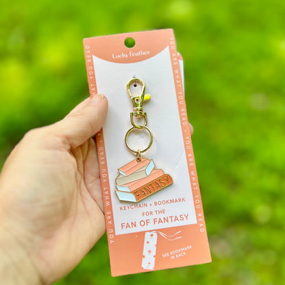 A hand holds a book-themed keychain and bookmark set on a peach display card against a blurred green outdoor background. The keychain features stacked books with the word 'FANTASY' in gold lettering, attached to a gold clasp.