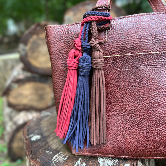 A close-up of a brown pebbled leather tote bag with braided handle straps and three hanging tassels in red, navy blue, and brown, resting on a rustic wooden log outdoors.