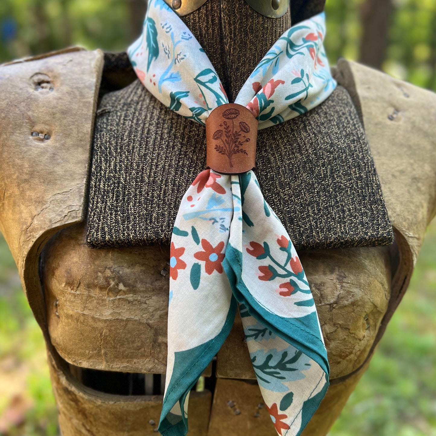 A floral neck scarf with a brown leather keeper, displayed on a medieval-style wooden armor chest piece against a blurred outdoor background.