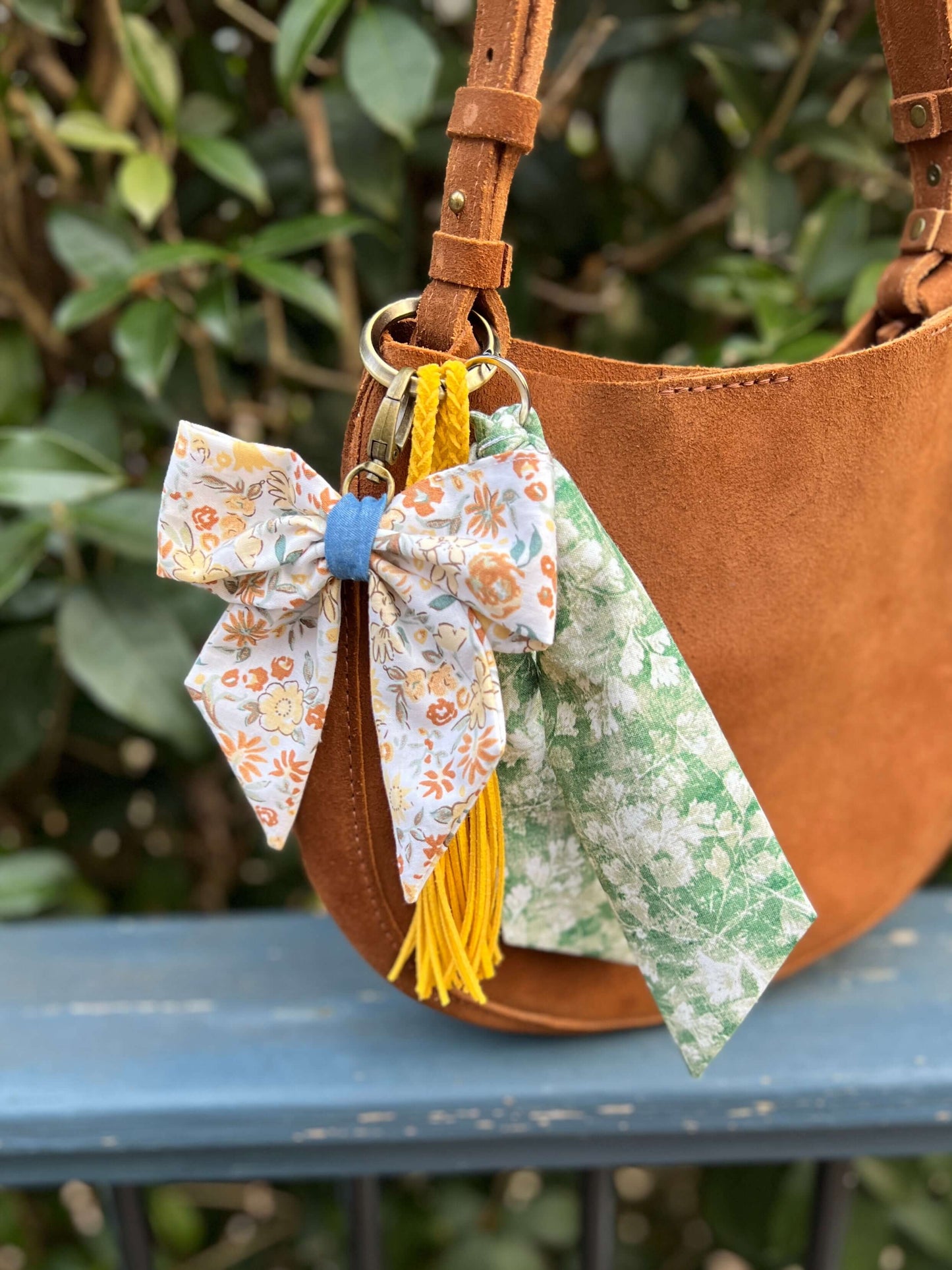 Brown handbag with floral bow keychain and tassels on a metal surface.