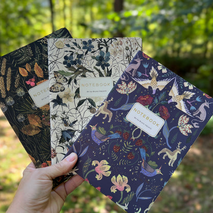 Three illustrated notebooks are fanned out in a hand outdoors, with a blurred forest background. The notebooks feature detailed botanical and fantasy illustrations in dark, earthy, and deep blue tones.