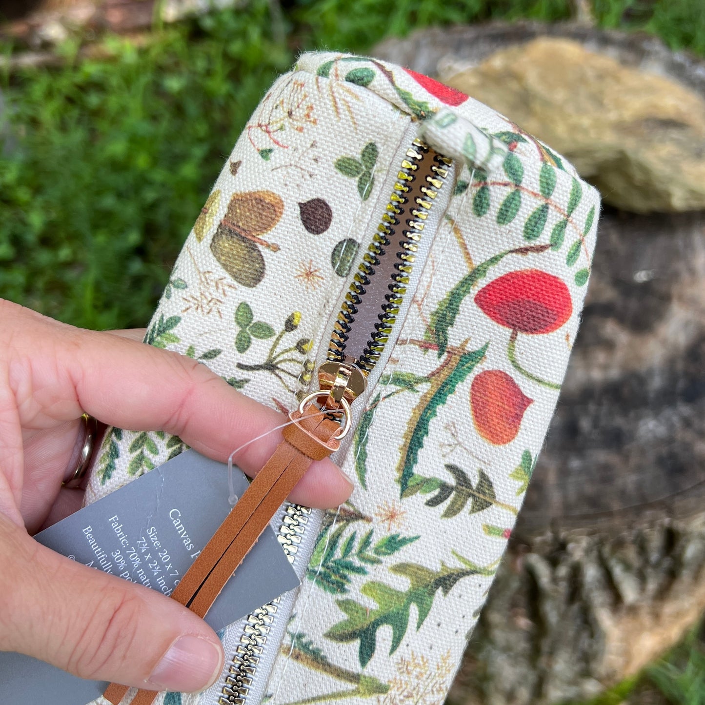 A close-up of a hand holding a canvas zip pouch with a botanical print featuring green leaves, red berries, and brown acorns. The pouch has a gold-tone zipper with a tan leather pull tab and a gray fabric hangtag.