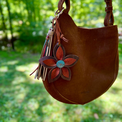 Brown leather handbag with a decorative leather flower bag charm against a blurred green outdoor background