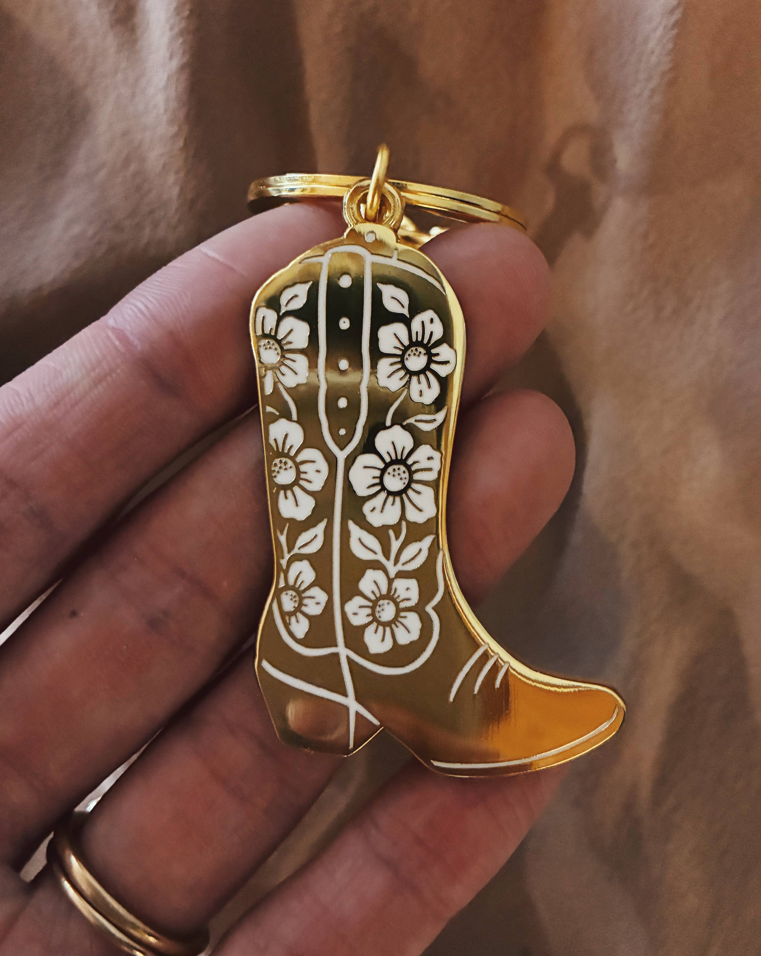 Gold keychain shaped like a cowboy boot with floral designs held in a hand against a brown background.