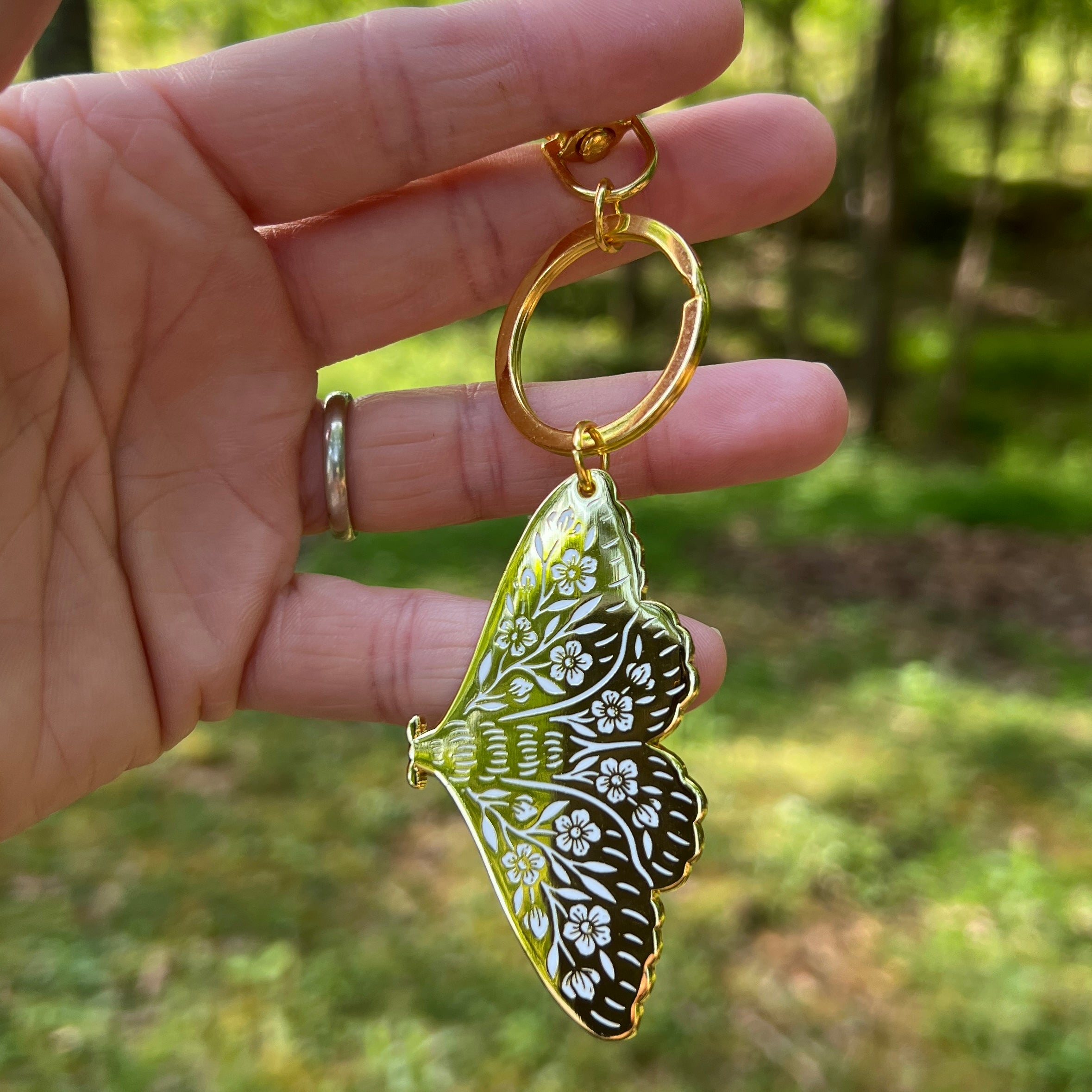 Hand holding a butterfly-shaped keychain with intricate design against a blurred natural background