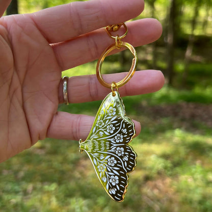 Hand holding a butterfly-shaped keychain with intricate design against a blurred natural background