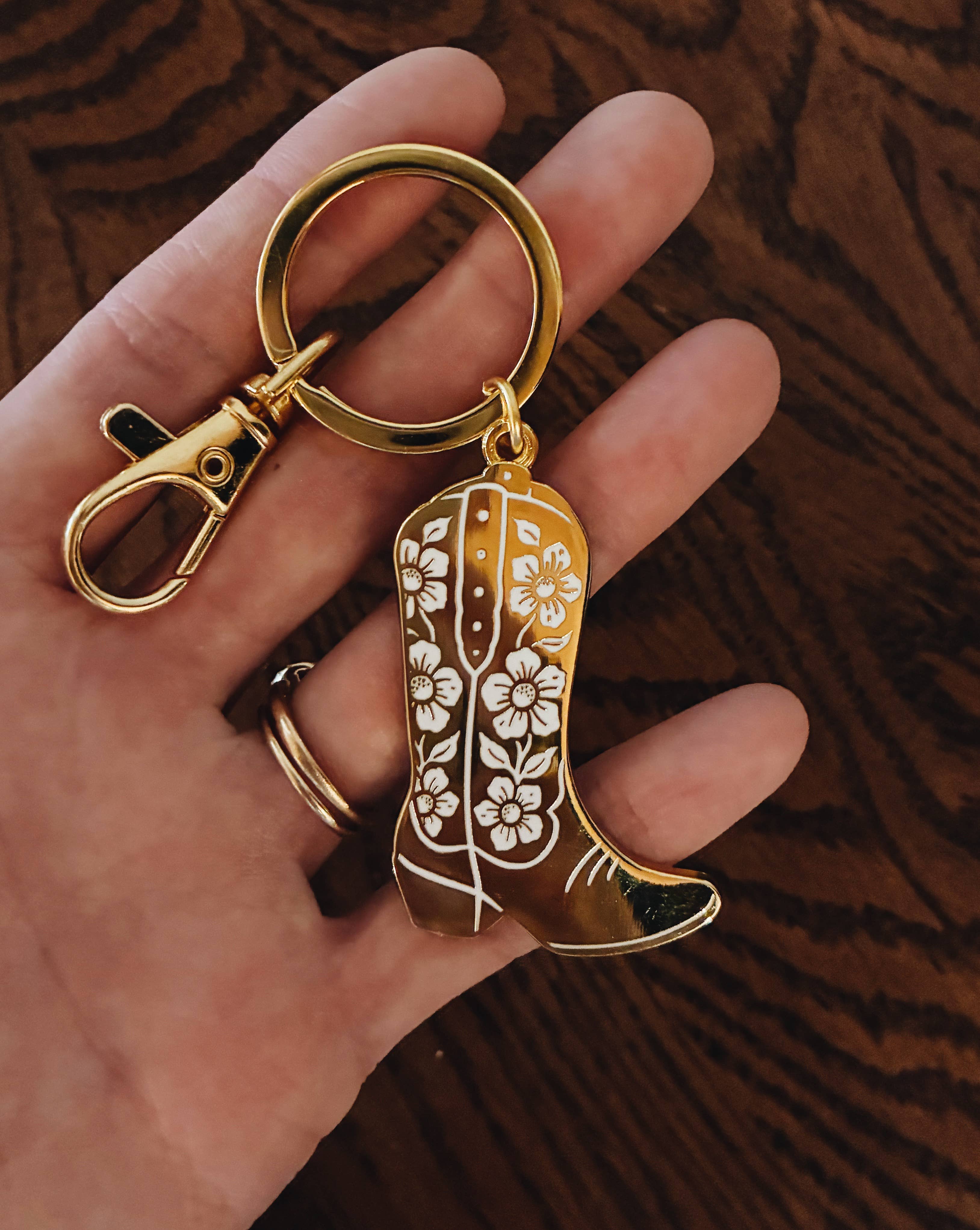 Gold keychain shaped like a boot with floral patterns held by a hand against a wooden background.
