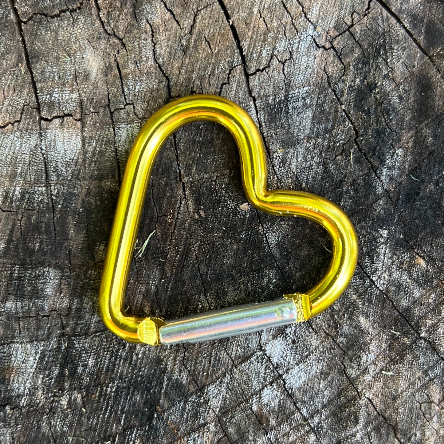 A heart-shaped metallic carabiner with a bright yellow body and a silver iridescent slide, resting on a textured, weathered wood surface.