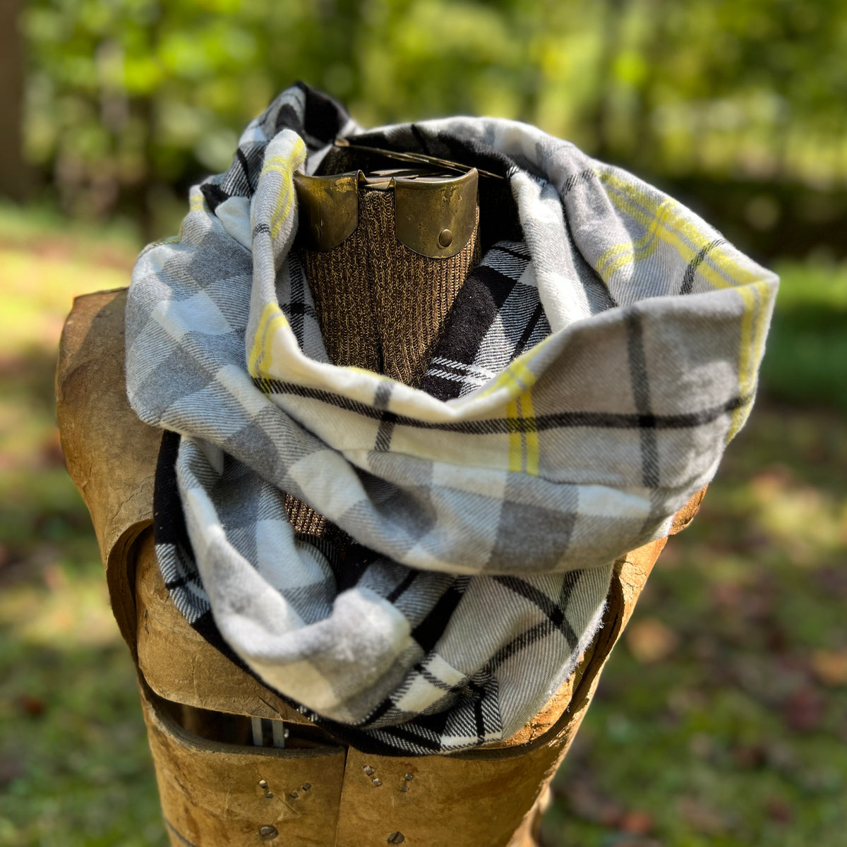 A plaid scarf in gray, white, black, and yellow is draped around a rustic wooden display stand outdoors with a blurred green forest background.