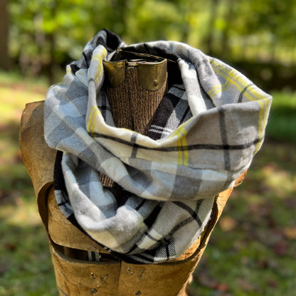 A plaid scarf in gray, white, black, and yellow is draped around a rustic wooden display stand outdoors with a blurred green forest background.