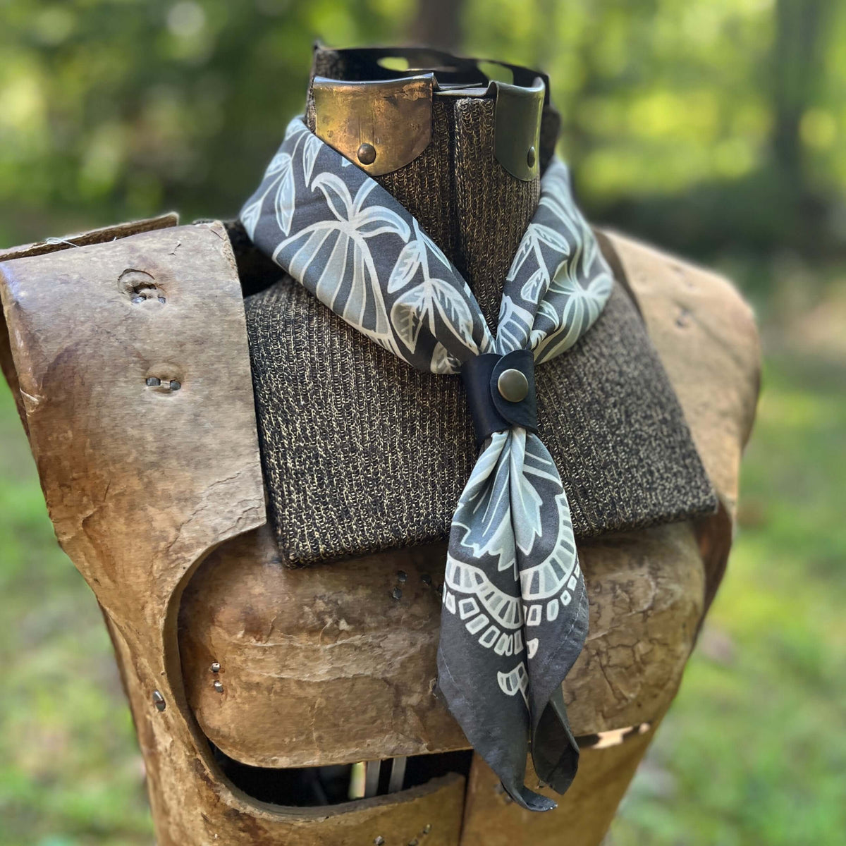 A patterned bandana-style neck scarf with gray, white, and black tropical leaf motifs, styled on a medieval wooden armor display outdoors with a blurred green forest background.