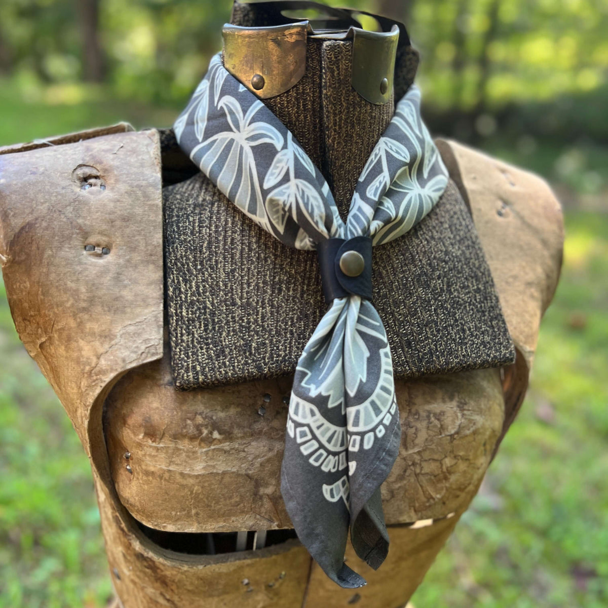 A gray and white tropical leaf print neck scarf styled on a rustic wooden armor display against a blurred green outdoor background.