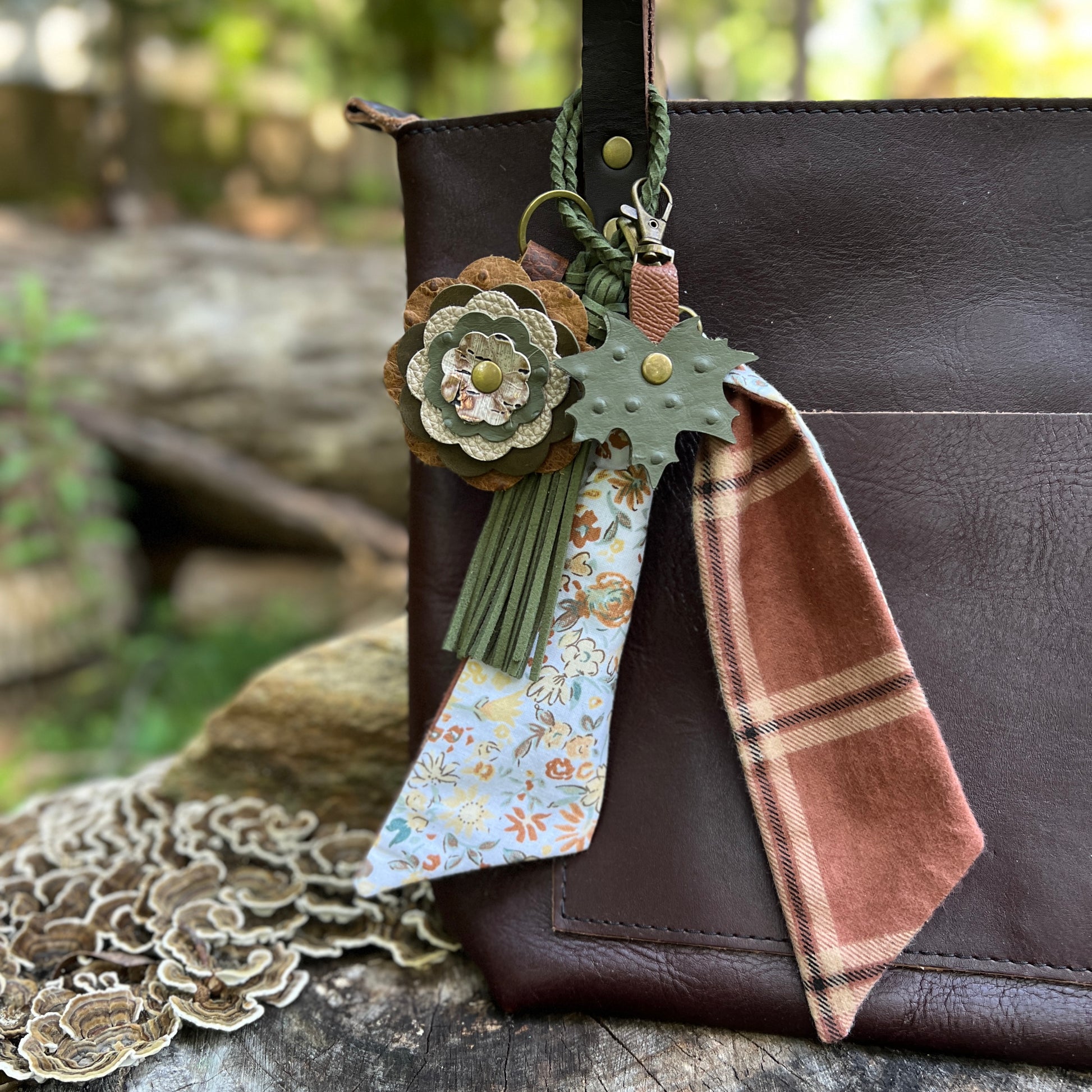 A brown leather shoulder bag with a decorative hanging keychain charm. The charm includes a green leaf-shaped tag, a rust-brown plaid fabric tag, green tassels, and a floral leather flower accent, shown against a blurred outdoor background.
