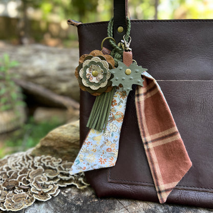 A brown leather shoulder bag with a decorative hanging keychain charm. The charm includes a green leaf-shaped tag, a rust-brown plaid fabric tag, green tassels, and a floral leather flower accent, shown against a blurred outdoor background.