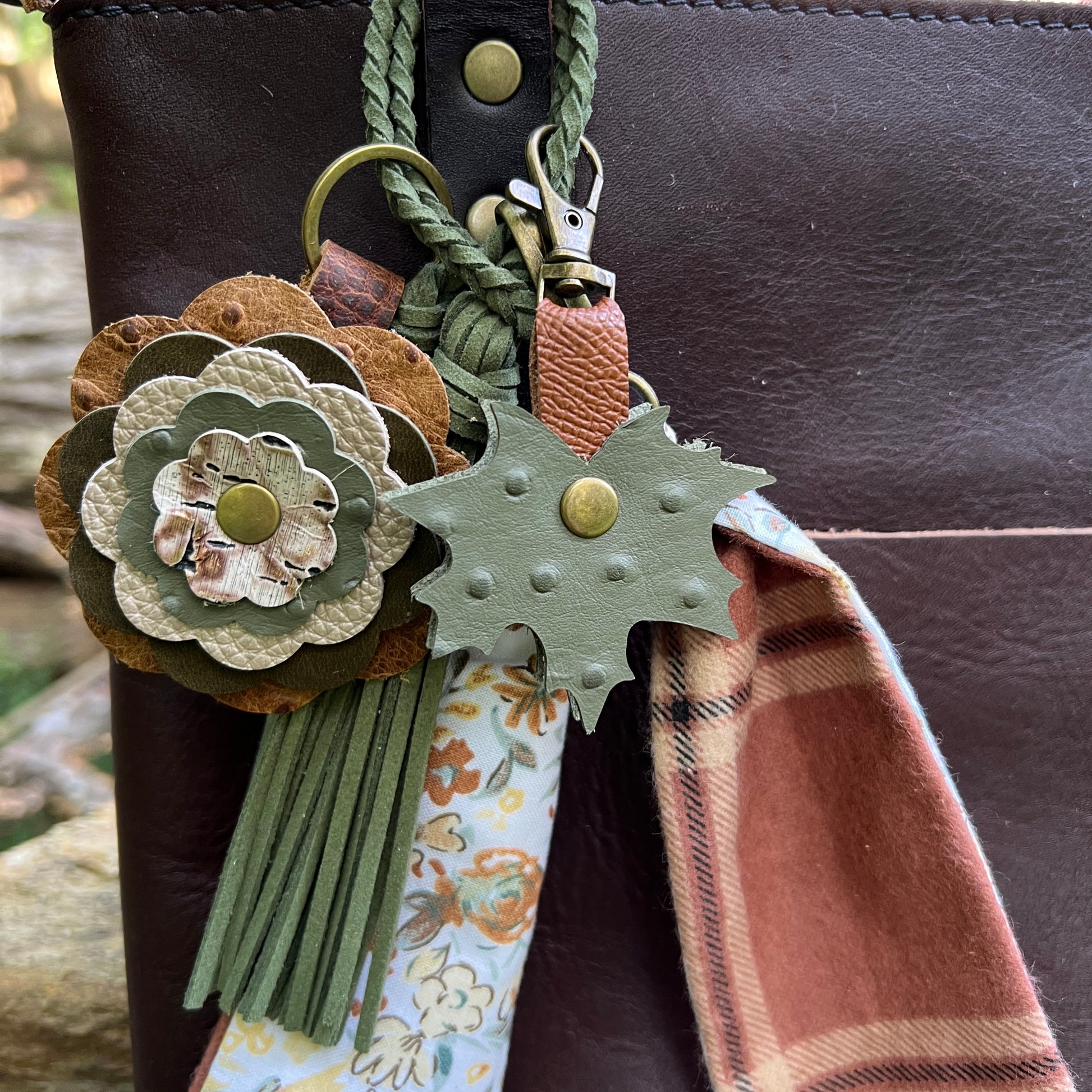 A close-up of a leather keychain or bag charm set attached to a dark brown leather bag. The set includes a layered flower-shaped leather ornament in tan, olive green, and cream, a green leaf-shaped ornament with brass studs, a green braided strap, a floral fabric tag, and a rust-brown plaid fabric accent.