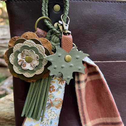 A close-up of a leather keychain or bag charm set attached to a dark brown leather bag. The set includes a layered flower-shaped leather ornament in tan, olive green, and cream, a green leaf-shaped ornament with brass studs, a green braided strap, a floral fabric tag, and a rust-brown plaid fabric accent.