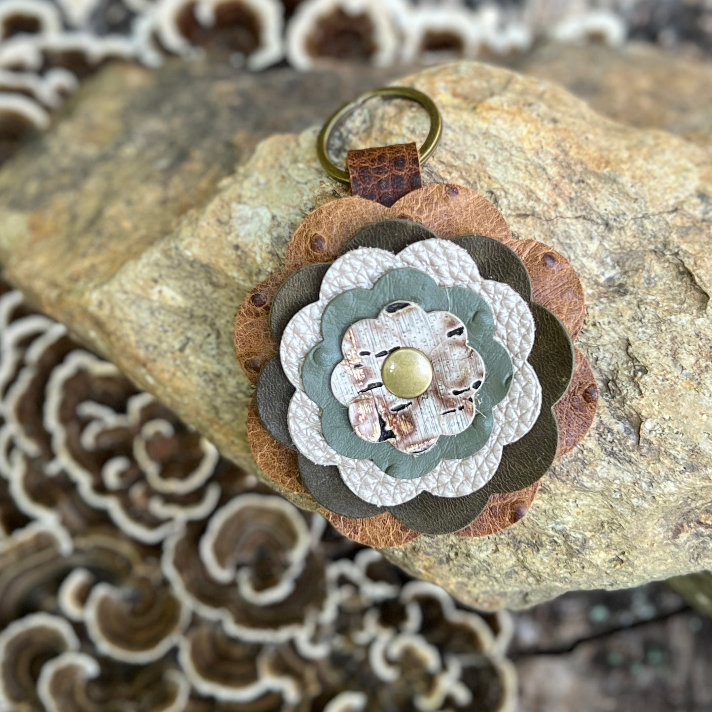 A handmade multi-layered flower keychain made from brown, sage green, and beige leather, featuring a small gold button in the center and a brass key ring, displayed on a rock with mushrooms in the background.