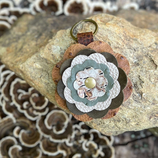 A handmade multi-layered flower keychain made from brown, sage green, and beige leather, featuring a small gold button in the center and a brass key ring, displayed on a rock with mushrooms in the background.