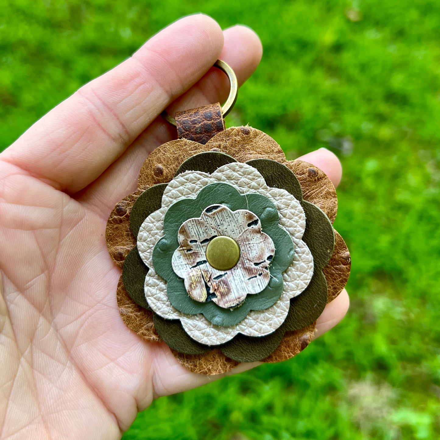 A handmade flower-shaped keychain or bag charm held in a hand, featuring layered brown, olive green, and cream textured leather petals with a small brass snap button at the center, set against a blurred green grass background.