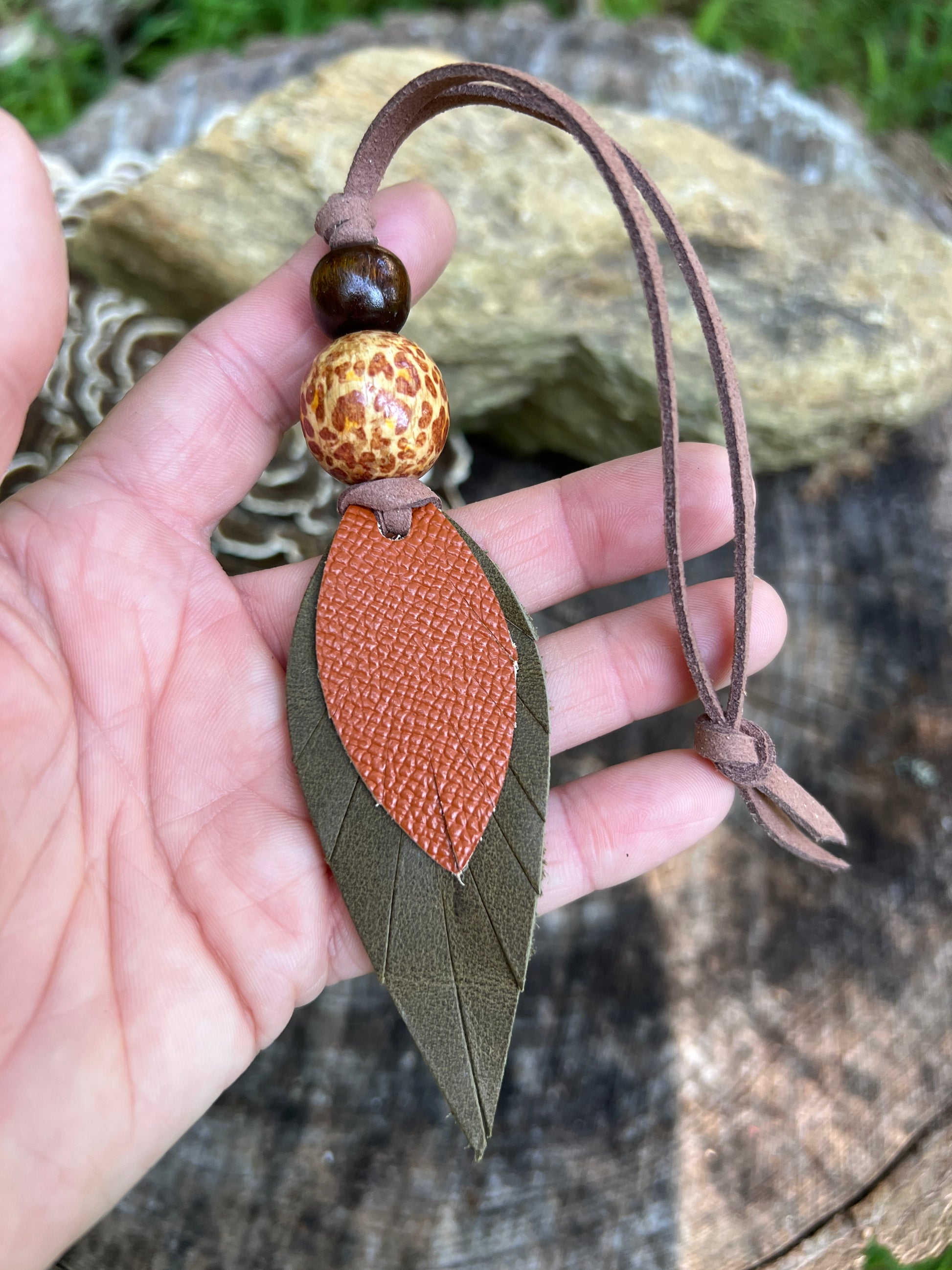 A hand holding a leaf-shaped leather bag charm with an olive green outer layer and a textured orange-brown center, attached to a brown leather cord with two decorative beads, against an outdoor background of rocks and wood.