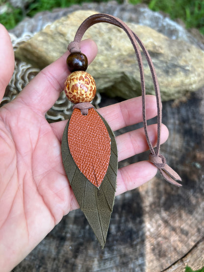 A hand holding a leaf-shaped leather bag charm with an olive green outer layer and a textured orange-brown center, attached to a brown leather cord with two decorative beads, against an outdoor background of rocks and wood.