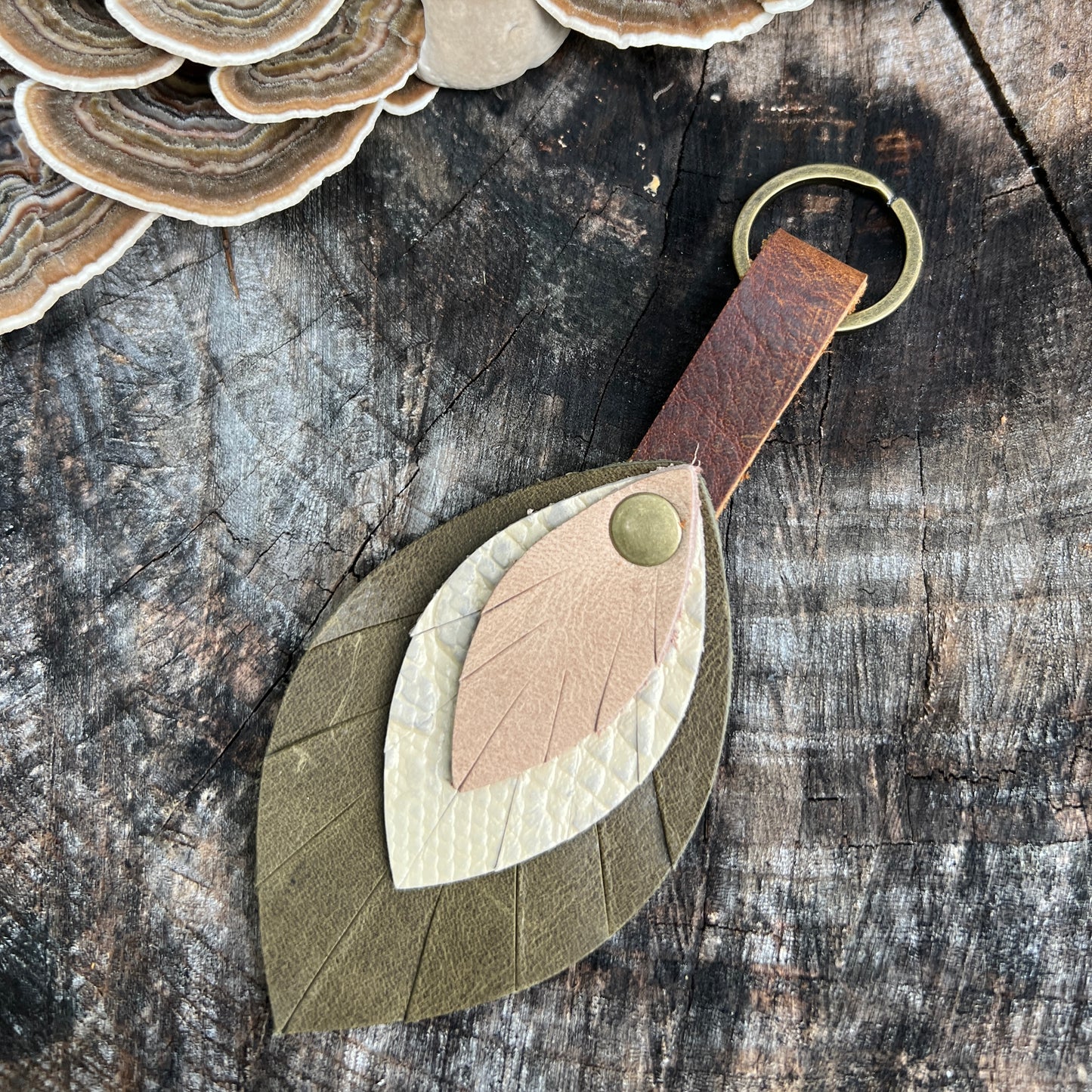 A leaf-shaped leather keychain or bag charm with layered natural leather pieces in olive green, cream, and tan, attached to a brown leather loop and brass key ring, displayed on a rustic wood surface with mushrooms in the corner.