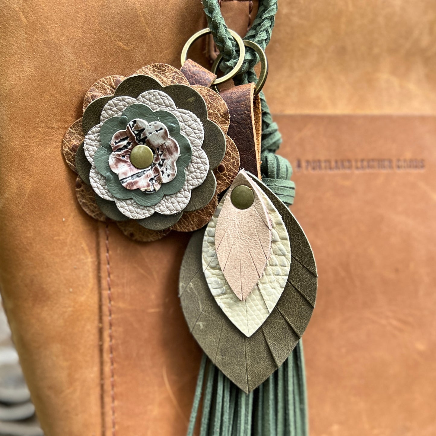 A close-up of a handmade leather keychain or bag charm with a braided green cord, a layered flower motif, a leaf-shaped charm, and a tassel, shown against a tan leather background.