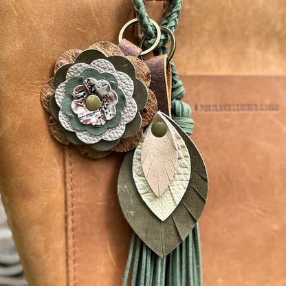 A close-up of a handmade leather keychain or bag charm with a braided green cord, a layered flower motif, a leaf-shaped charm, and a tassel, shown against a tan leather background.