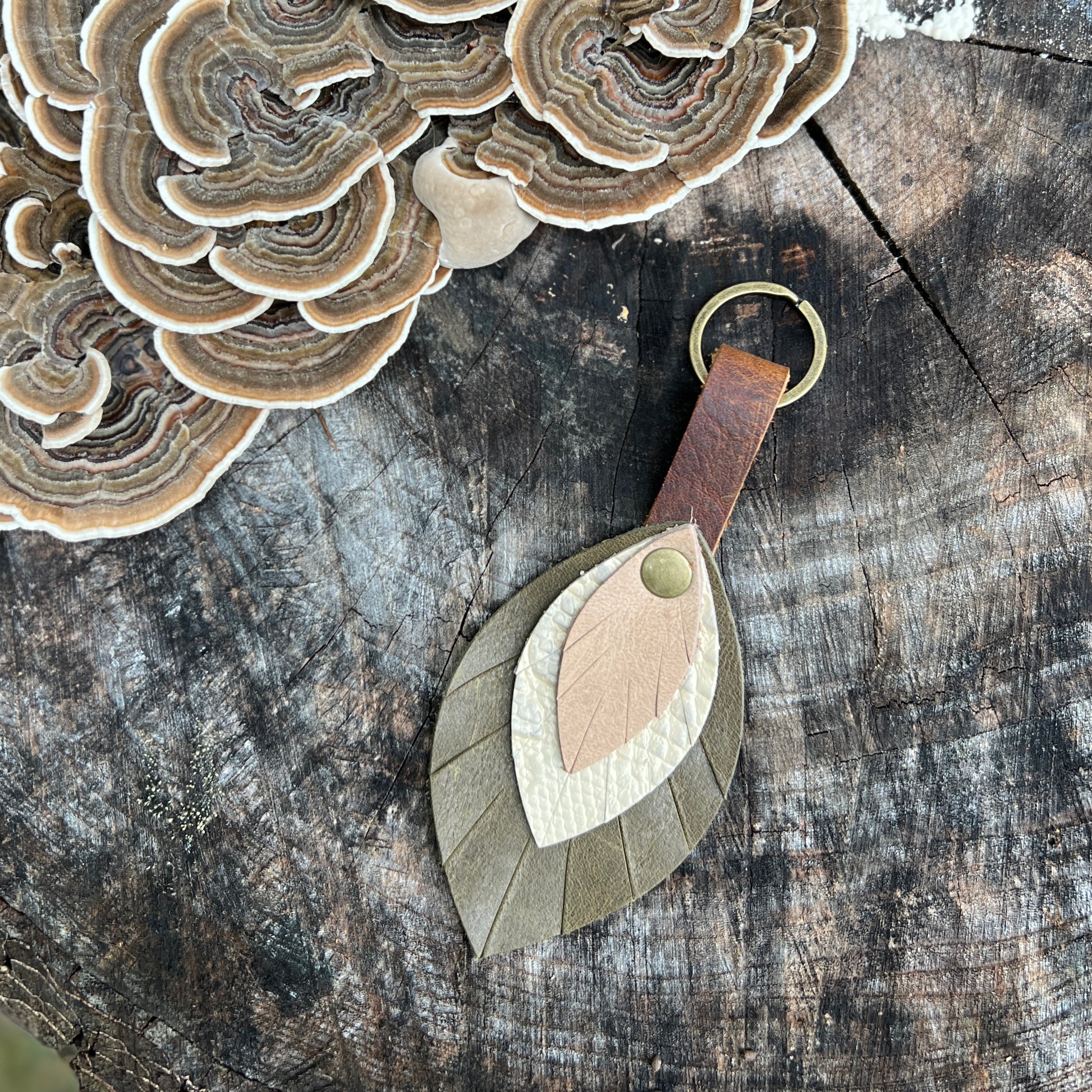A leaf-shaped leather keychain with layered tan, cream, and olive green pieces, a brown leather loop, and a brass key ring, displayed on a rustic wood surface next to shelf fungi.