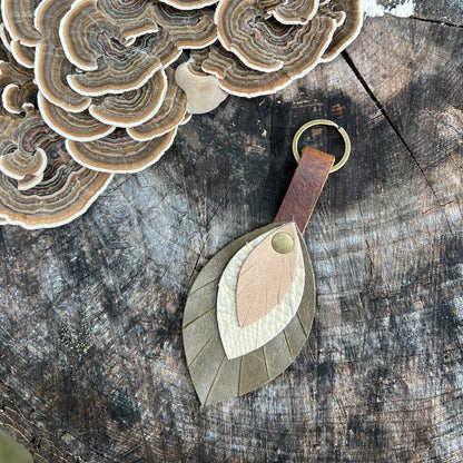 A leaf-shaped leather keychain with layered tan, cream, and olive green pieces, a brown leather loop, and a brass key ring, displayed on a rustic wood surface next to shelf fungi.