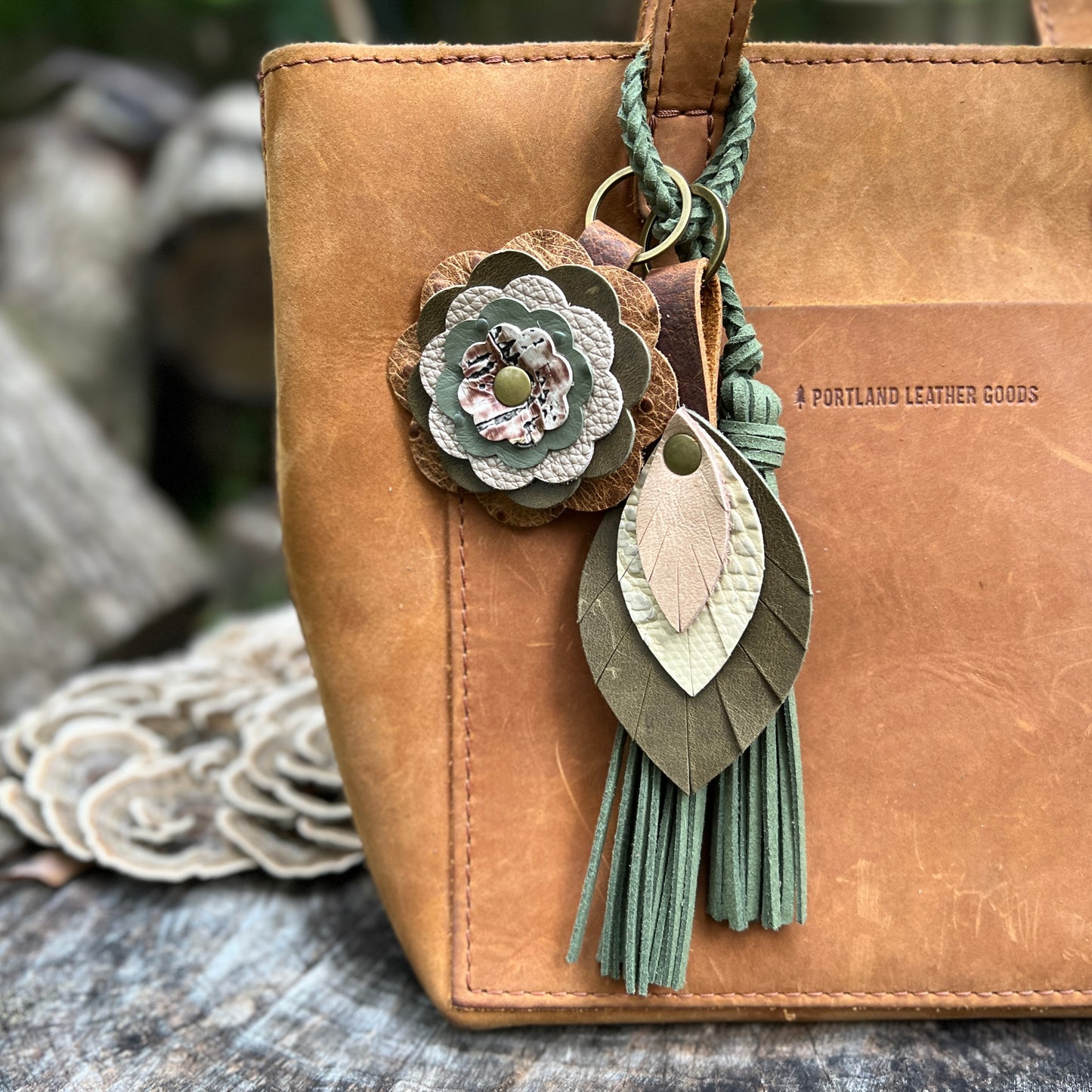 A tan leather tote bag with a decorative hanging keychain charm. The charm includes a layered flower, a braided green cord, and two leaf-shaped leather pieces with fringe tassels.