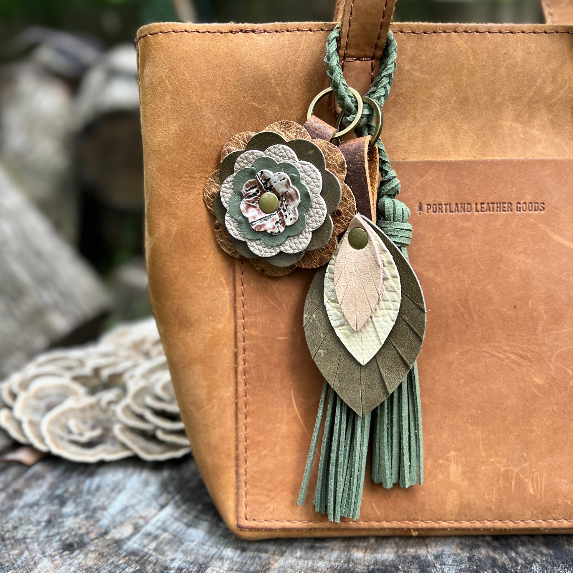 A tan leather tote bag with a decorative hanging keychain charm. The charm includes a layered flower, a braided green cord, and two leaf-shaped leather pieces with fringe tassels.