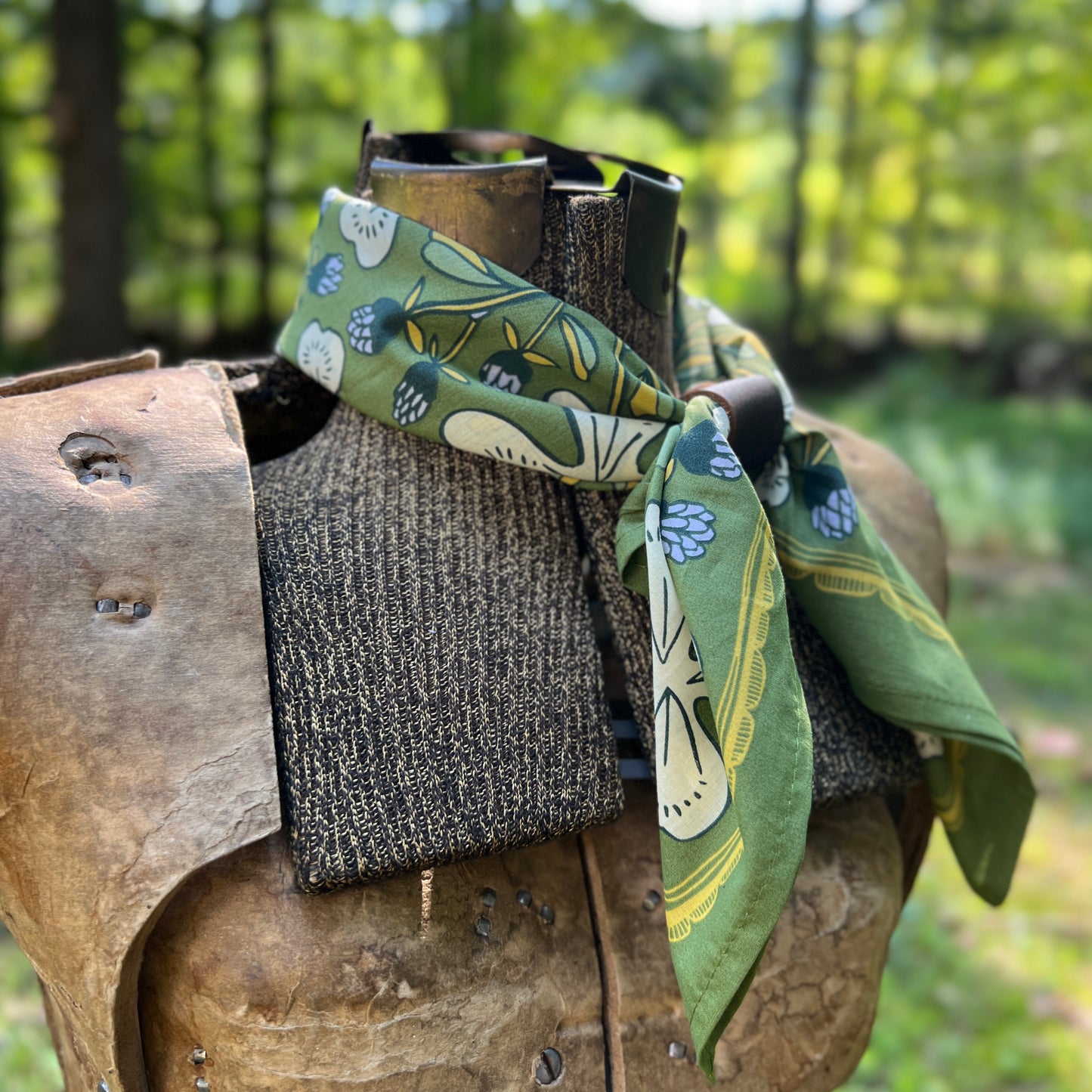 A green patterned cotton scarf with blue and cream floral motifs, tied around a rustic wooden display stand outdoors with a blurred forest background.