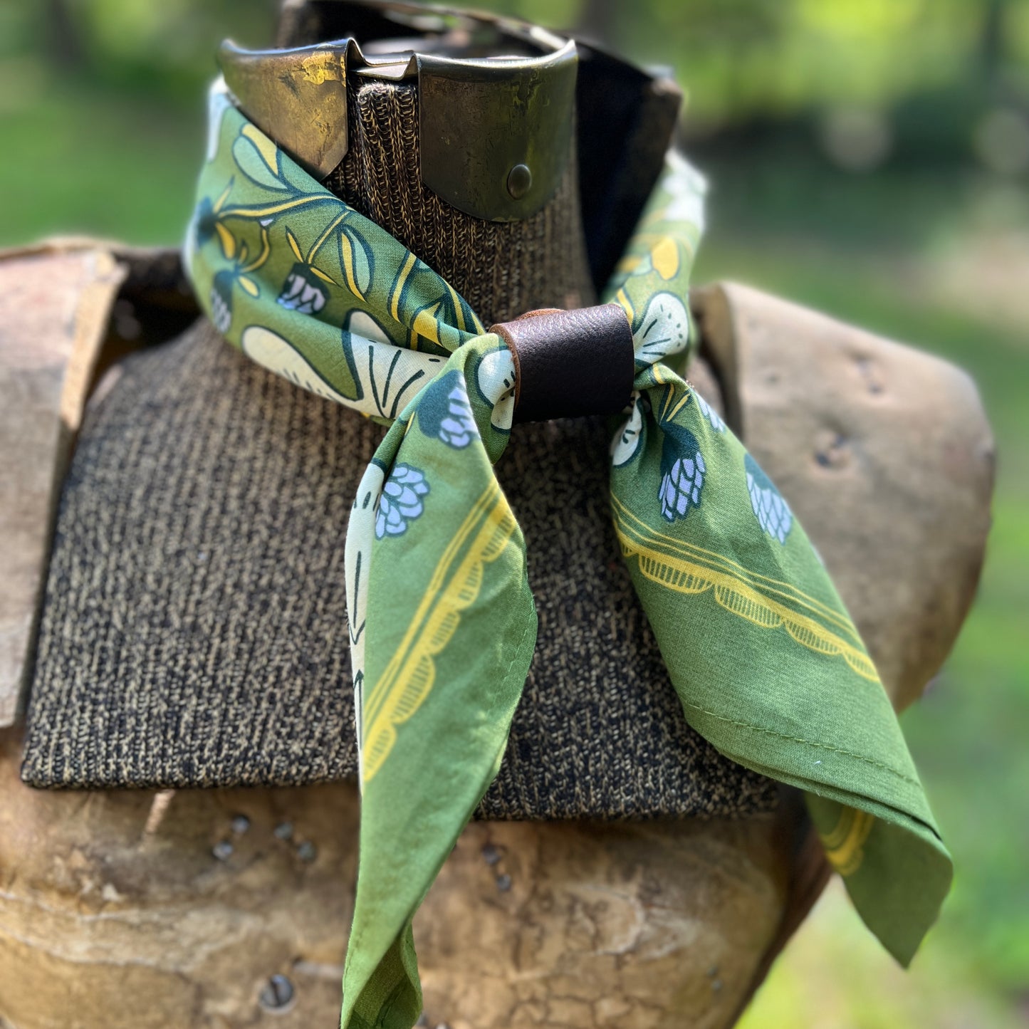 A green patterned neck scarf with a dark brown leather wrap, styled on a rustic wooden armor display against a blurred green outdoor background.