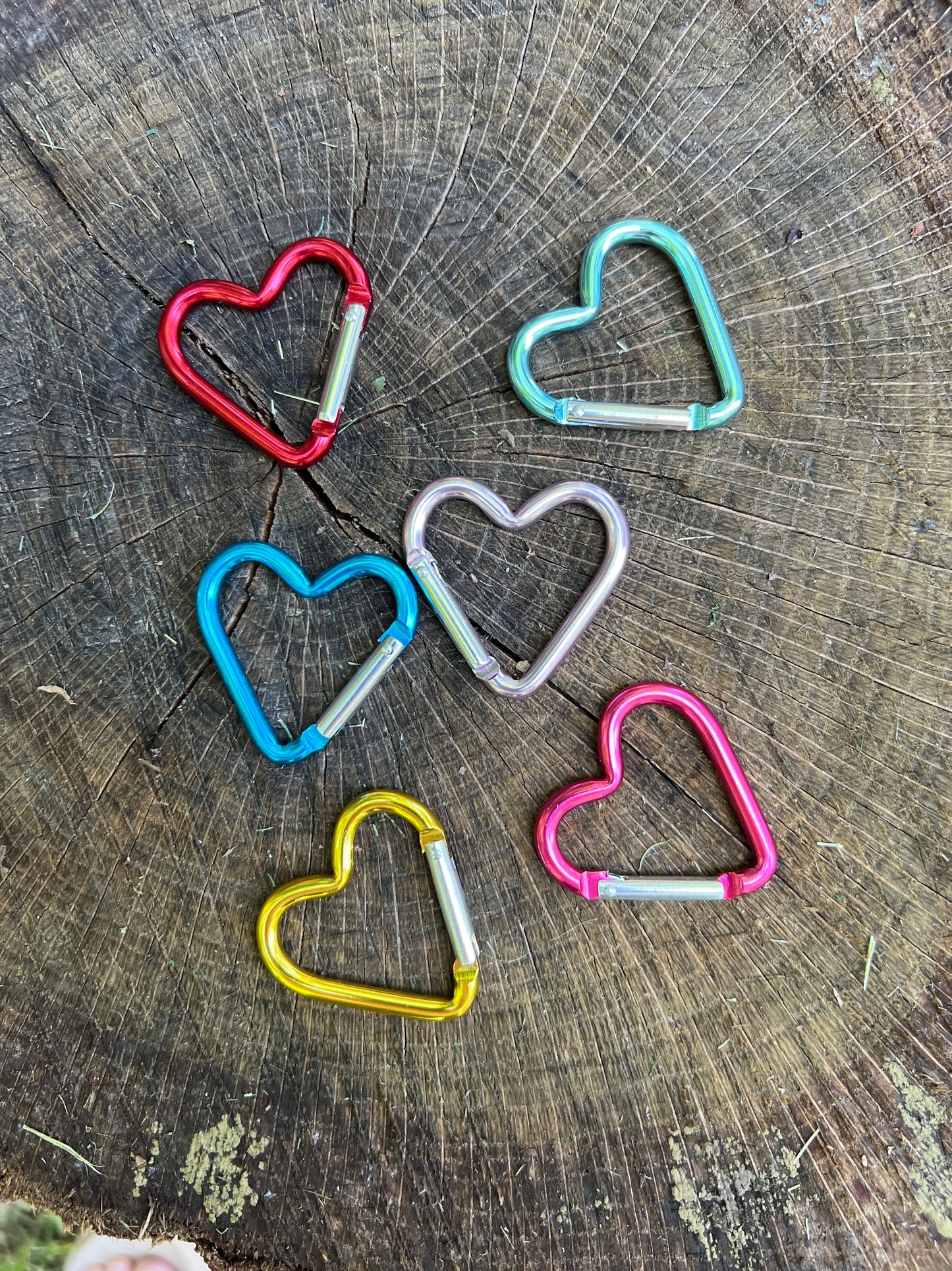 Five heart-shaped metal carabiners in red, turquoise, blue, silver, yellow, and pink, arranged on a rustic wood log background.