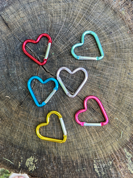 Five heart-shaped metal carabiners in red, turquoise, blue, silver, yellow, and pink, arranged on a rustic wood log background.