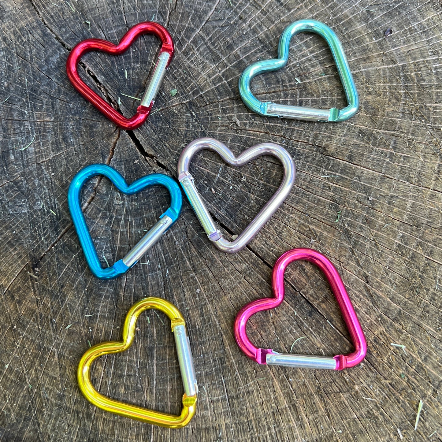 Five heart-shaped metal carabiners in red, mint green, blue, silver, and pink, arranged on a textured wood surface.