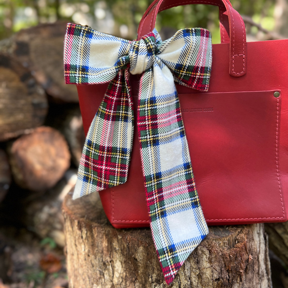 A red leather tote bag with a large plaid bow in cream, red, green, and blue, resting on a rustic tree stump with stacked logs in the background.