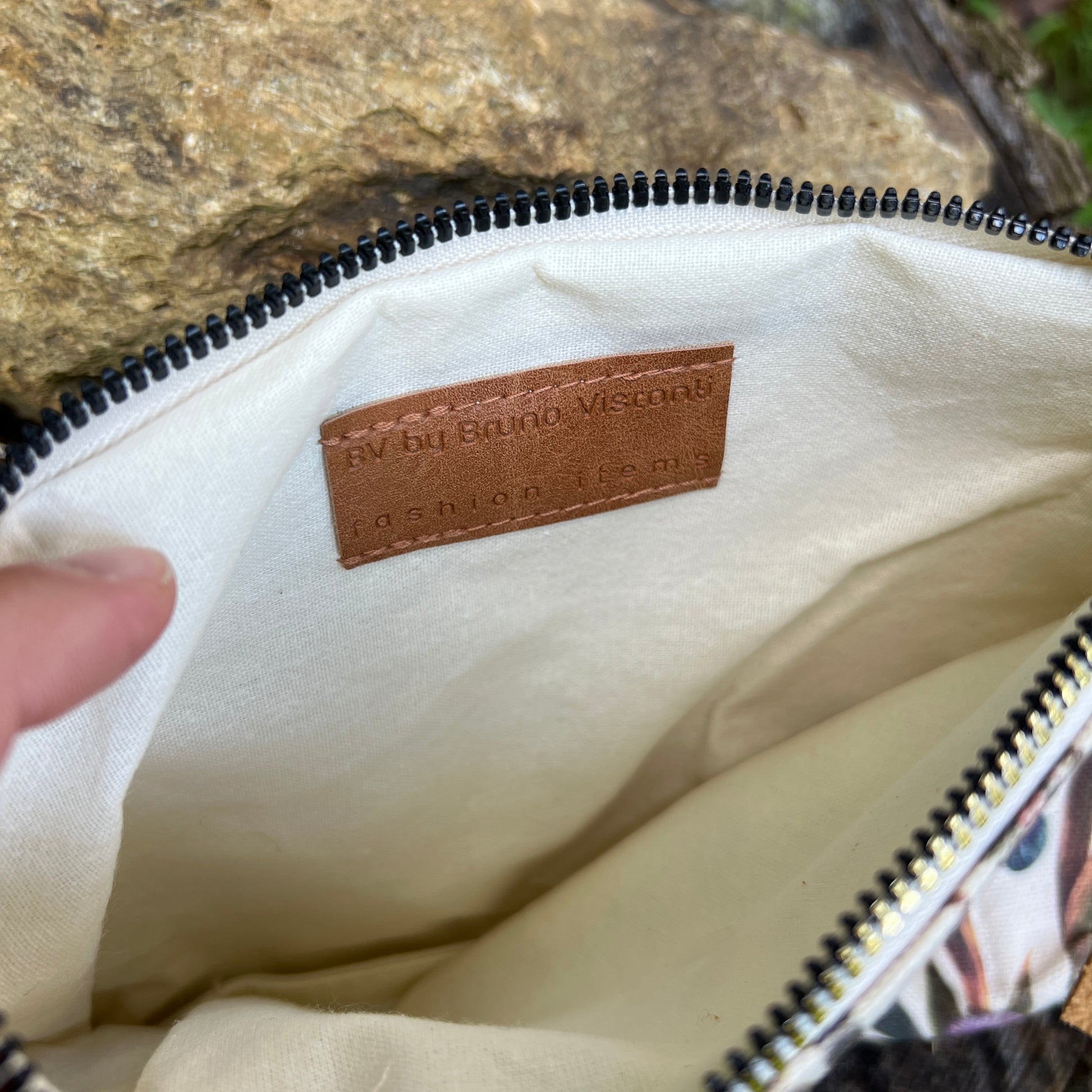 Close-up of the interior of a zippered pouch or bag, showing a cream canvas lining and a brown leather brand label that reads 'by by Bordo Visconti fashion leather.' A finger is visible at the left edge holding the opening open, with a rock in the background.