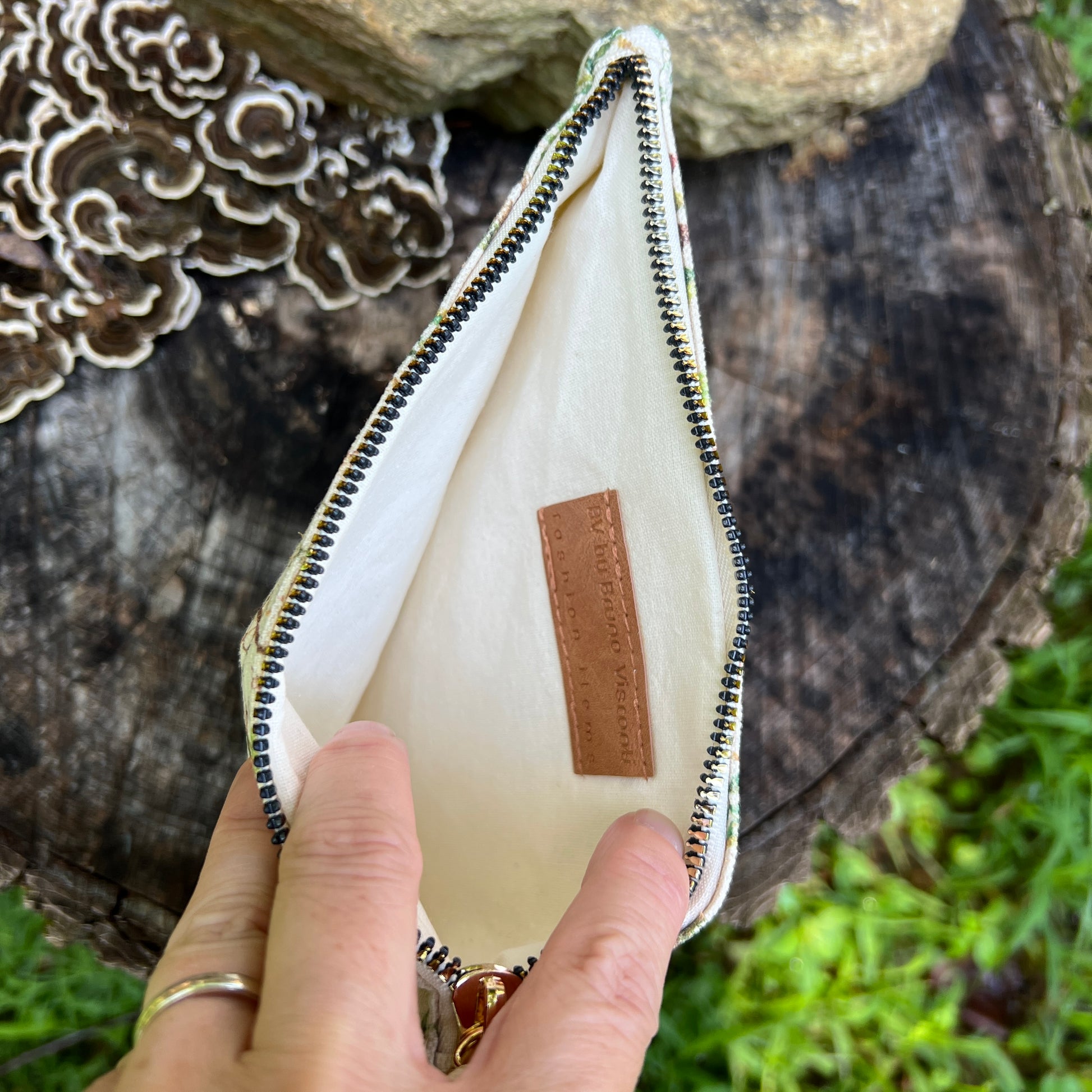 A hand holding open a small zip pouch with a cream interior and a tan brand patch reading 'The Art of Traveling'. The pouch is photographed outdoors on a tree stump with mushrooms and greenery in the background.