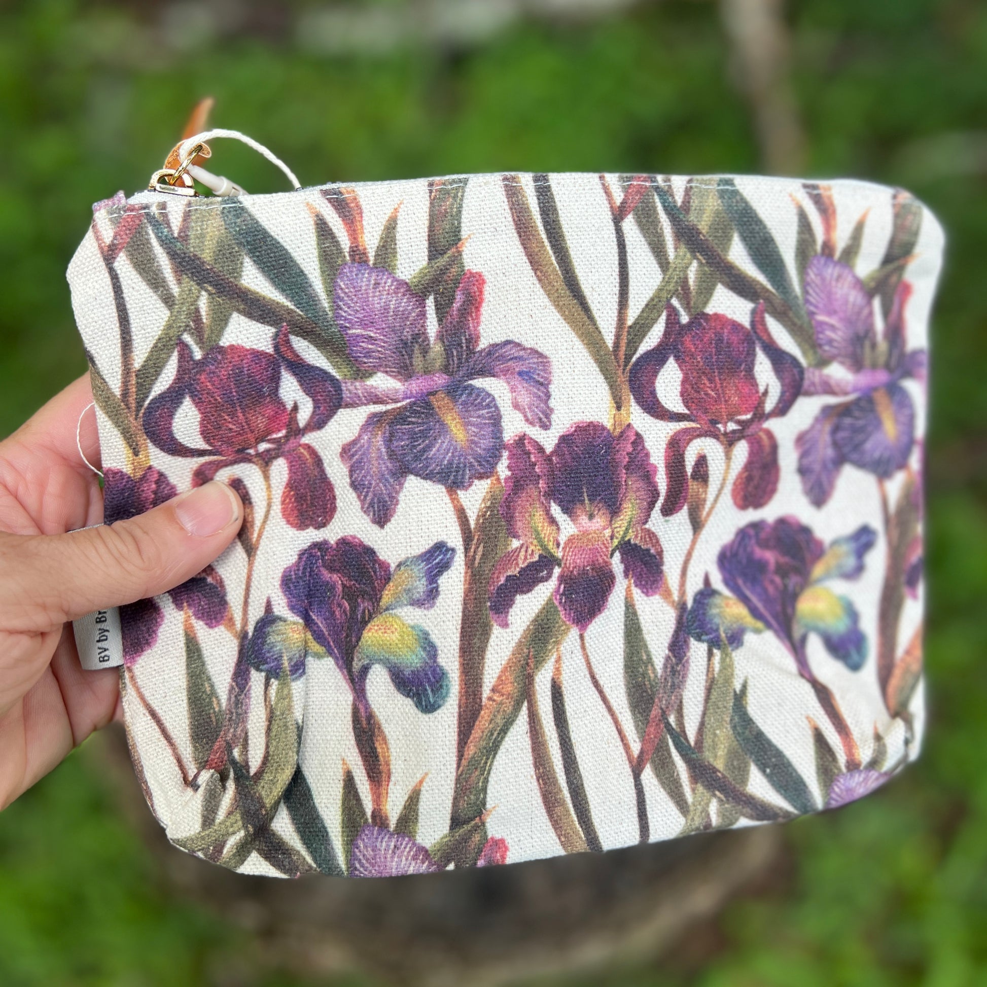 A hand holds a zippered pouch featuring a detailed print of purple irises on a white background, with a blurred green outdoor environment behind it.