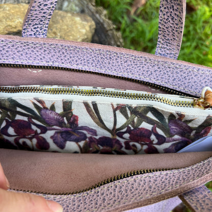 Close-up of a lavender pebbled leather handbag showing the interior lining with a purple and green floral print and a gold zipper.
