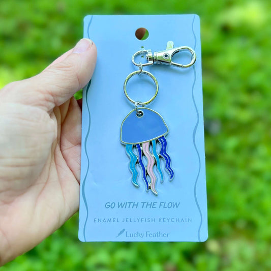 An enamel jellyfish keychain with a silver-tone lobster clasp, displayed on its light blue backing card held by a hand against a blurred green outdoor background. The backing card features the brand name 'Lucky Feather' and the text 'GO WITH THE FLOW'.