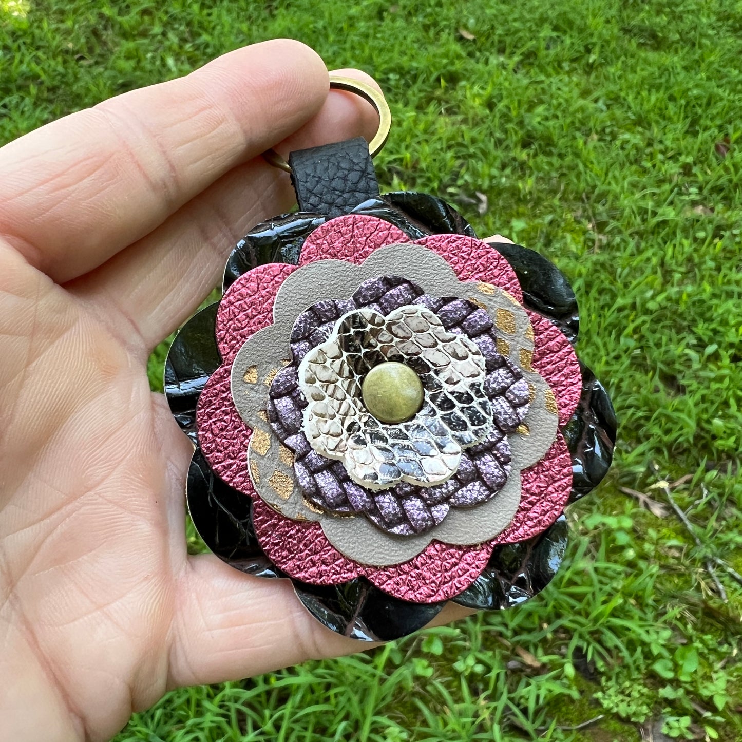 A hand holding a decorative flower-shaped keychain or bag charm outdoors against a green grass background. The charm features multiple textured leather and snakeskin-patterned flower layers in pink, black, beige, and purple, with a small gold-tone metal ring at the top and a central gold bead.