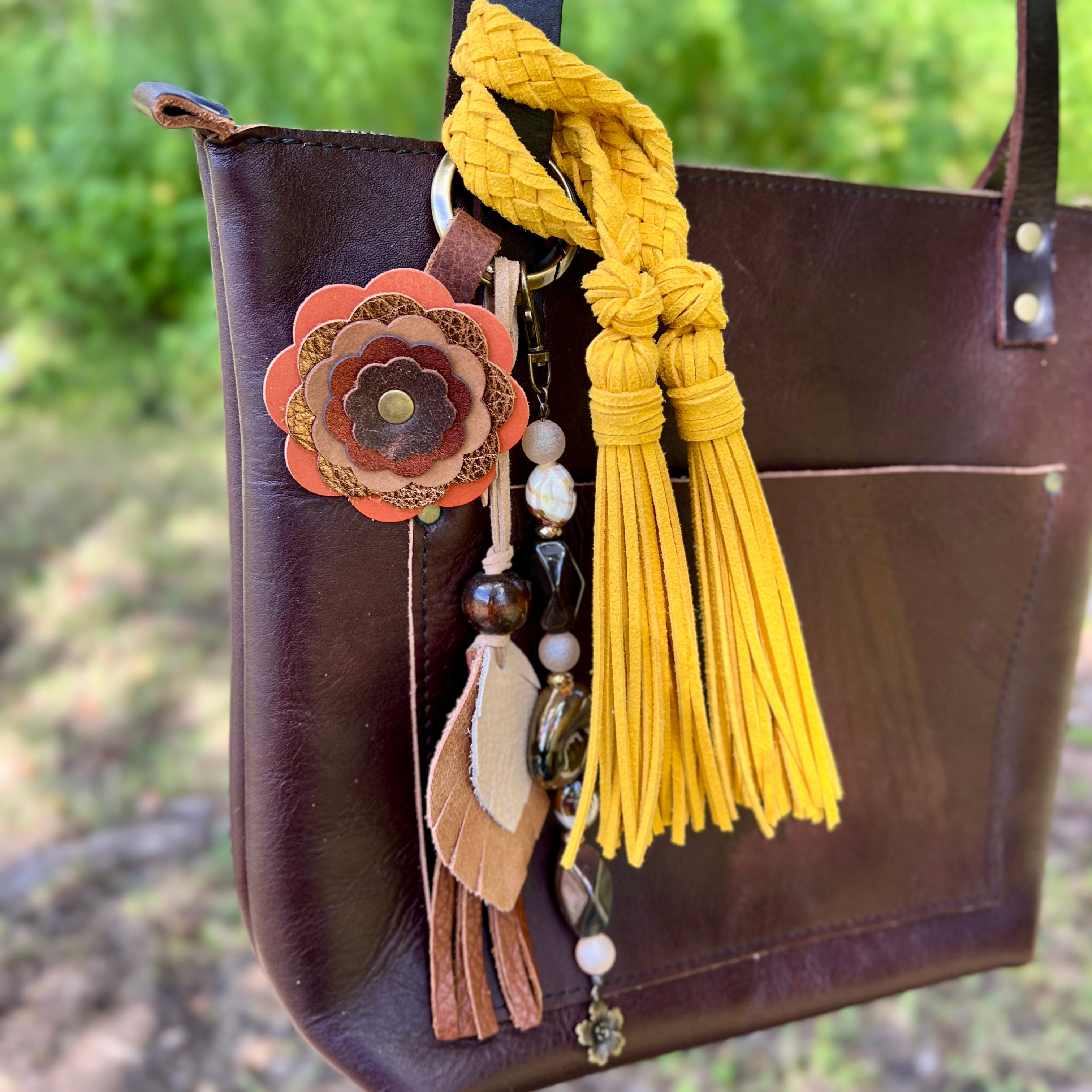 Brown leather handbag with decorative tassel and floral charm against a blurred natural background